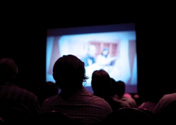 An unknown number of people sitting in a dark theater.  There is a projector screen in front of them with a man and woman at a conference.