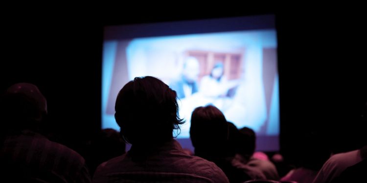 An unknown number of people sitting in a dark theater.  There is a projector screen in front of them with a man and woman at a conference.