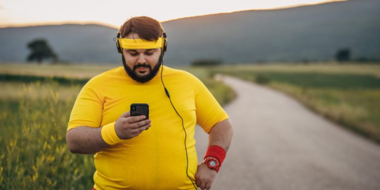 One man, young overweight man break after jogging, listening music on headphones, using smart phone.