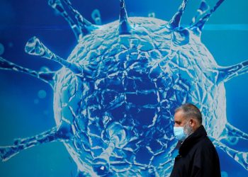A man wearing a protective face mask walks past an illustration of a virus (File: reuters.com)