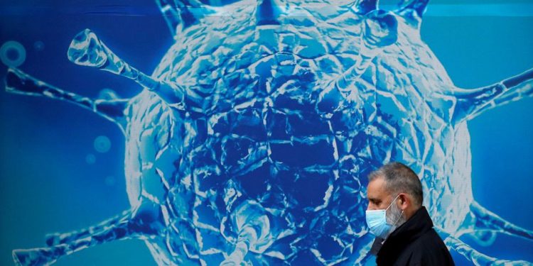 A man wearing a protective face mask walks past an illustration of a virus (File: reuters.com)