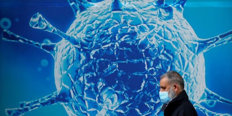 A man wearing a protective face mask walks past an illustration of a virus (File: reuters.com)